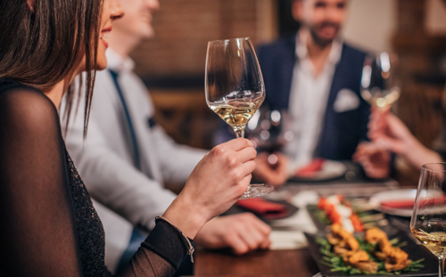 A woman holds a glass of wine whilst sitting at a table next to two men