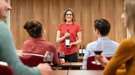 A woman stands in front of a class, holding a bottle of wine